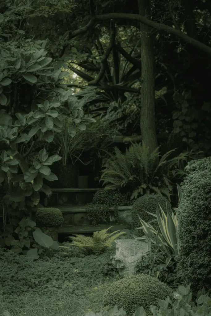 moody garden corner with shade and depth, dark green foliage, ferns and moss, stone elements, filtered light through trees, early morning atmosphere, quiet and mysterious mood, editorial garden photography, no people, no text, no logos
